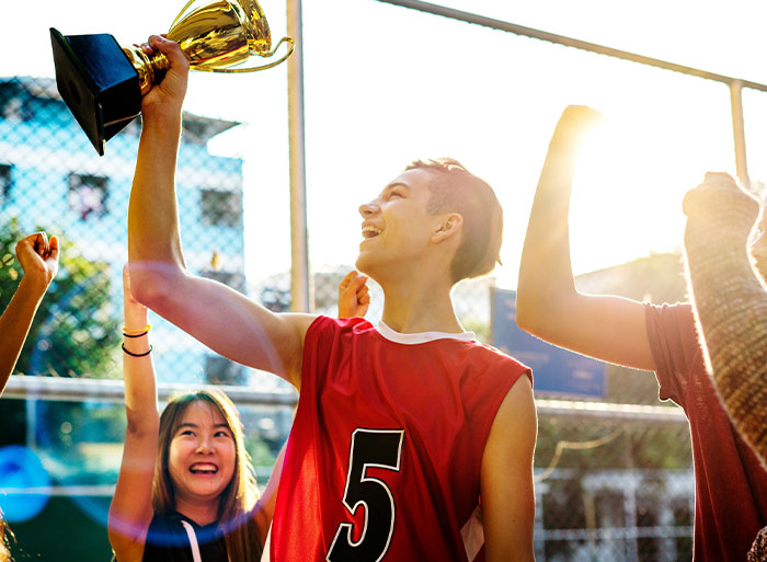 Teen in a basketball jersey holding a trophy, celebrating with friends under the sun.