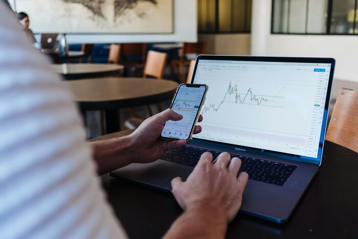 Person analyzing stock charts on laptop and phone in a modern workspace.