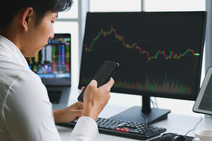 Person analyzing stock market graphs on a computer monitor and mobile phone.