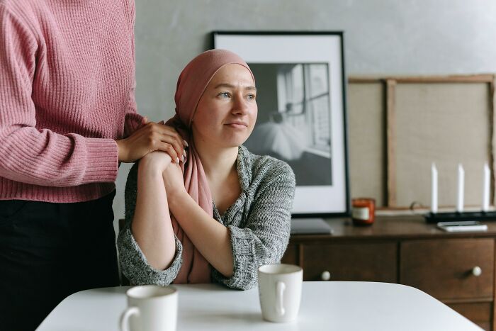 Person in a pink headscarf sitting at a table with two cups, receiving a supportive hand.