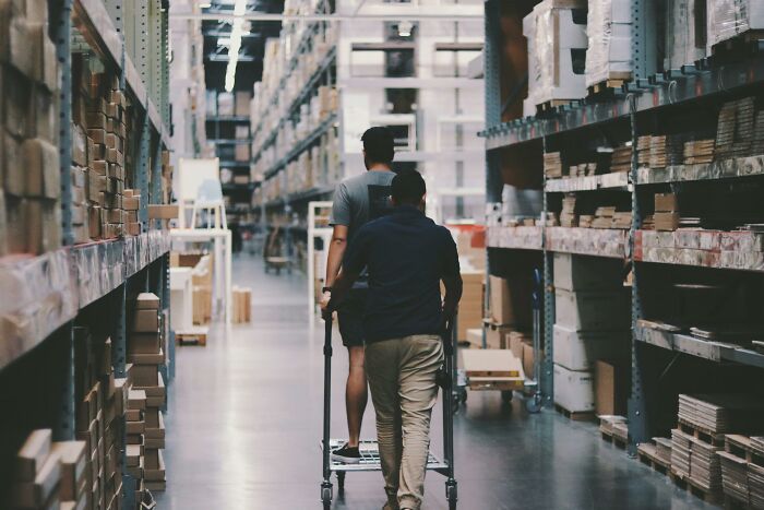 Two people walking through a warehouse aisle, one person pushing a cart.