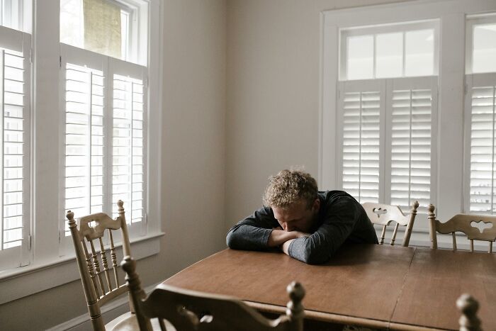 Man sitting at a wooden table with head down, appearing thoughtful and alone, next to a window with shutters, evoking secrets.