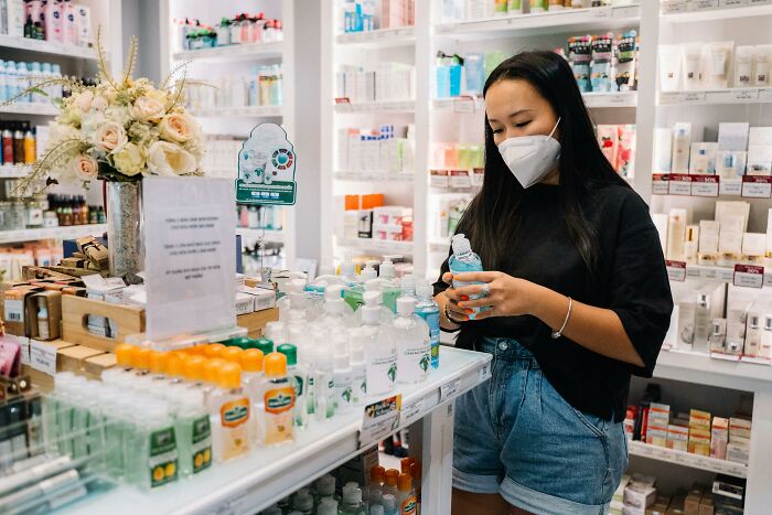 Woman in a black shirt and mask examines a product in a pharmacy, surrounded by shelves with various items, showcasing human genius.