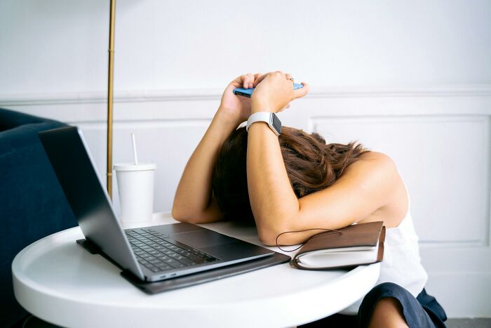 Millennial overwhelmed at a desk with a laptop, head resting on arms in a modern workspace.