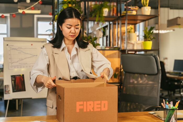 Millennial woman packing office belongings into a box labeled "fired," signaling challenges in adulthood transition.