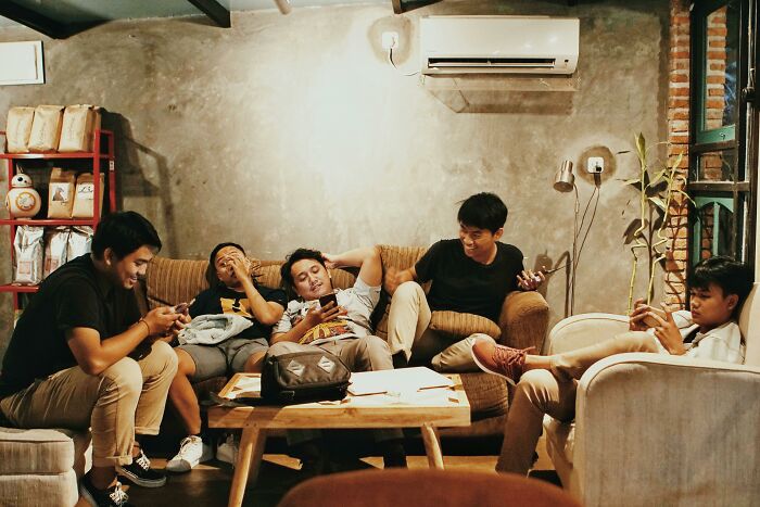 Group of young adults socializing in a cozy room, sitting on couches and using their phones.