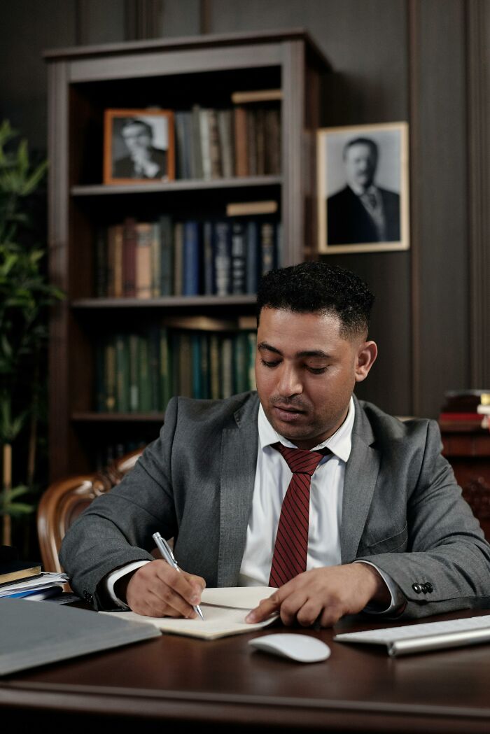 Man in a gray suit writing at a desk, surrounded by bookshelves.