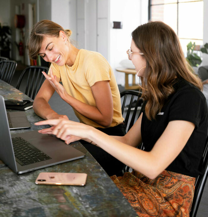 Two women laughing at a work desk, pointing at a laptop, symbolizing social norms interaction.