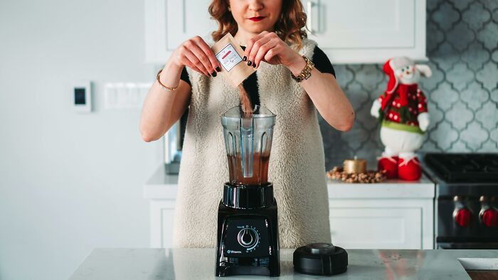 A woman using a high-end blender in a modern kitchen, pouring contents into the blender.