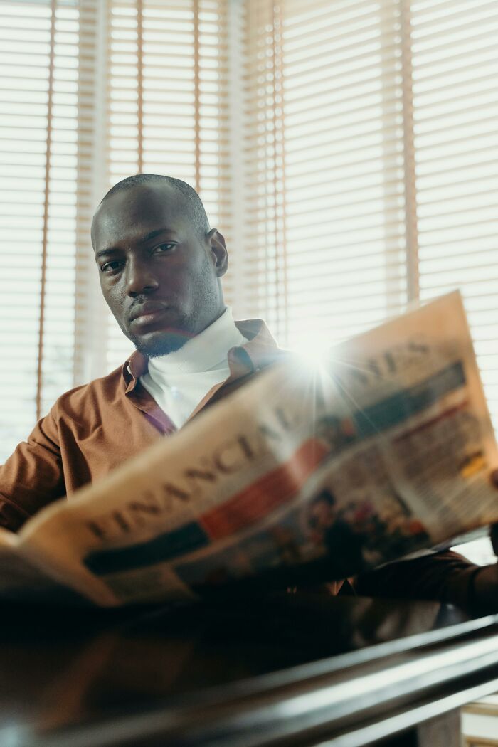 Man reading a financial newspaper with sunlight filtering through blinds.