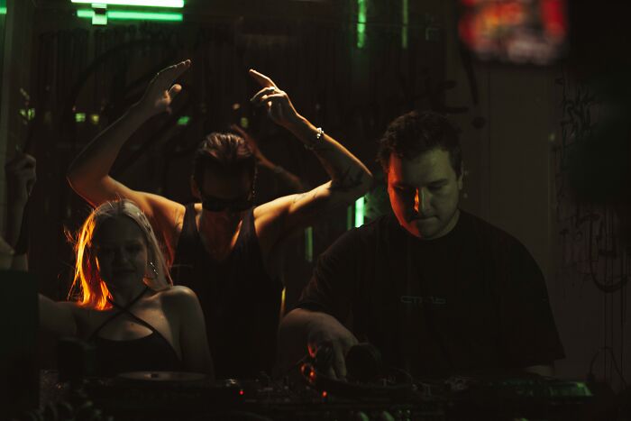 DJ performing music in a dark, eerie club scene with two people dancing, creating a creepy and mysterious atmosphere.