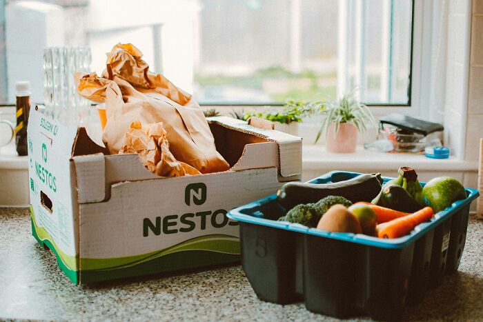 Box of groceries and fresh vegetables on a kitchen counter.