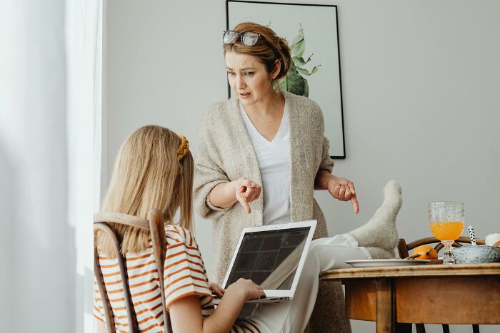 Woman pointing at a child using a laptop at a table, highlighting signs of a horrible parent.