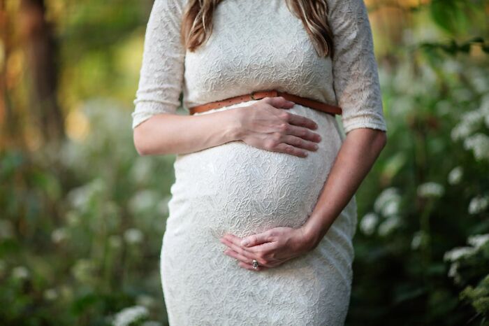 Pregnant woman in a lace dress in a garden, gently holding her belly, conveying intriguing life moments.