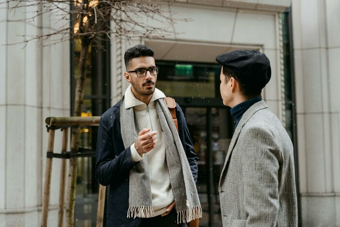 Two people socializing outdoors, one wearing glasses and a scarf, the other in a hat and a gray coat.