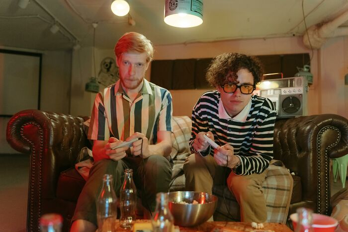 Two men sitting on a couch in a dimly lit room, holding game controllers, with a slightly unsettling atmosphere.