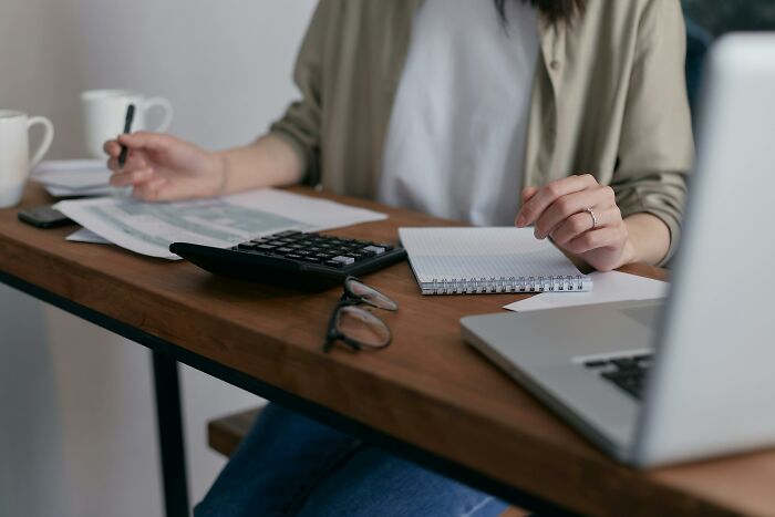 Person working at a desk with a calculator, notebook, and laptop, analyzing figures related to a financial scam.