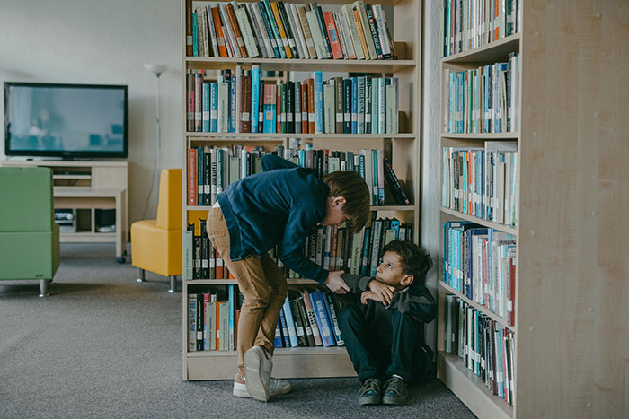 Boy being bullied in a library while sitting near a bookshelf.