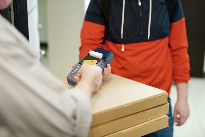 Delivery person hands over a card reader to a customer holding pizza boxes, emphasizing awkward delivery scenes.