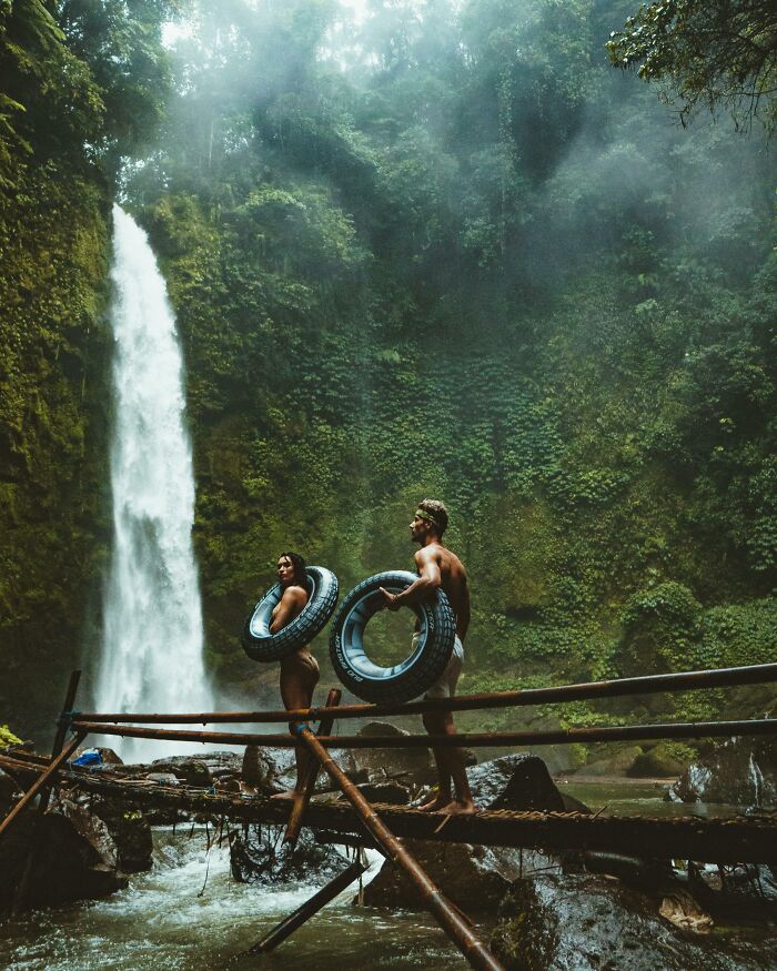 Two people holding inner tubes stand on a bridge near a waterfall in a lush forest.