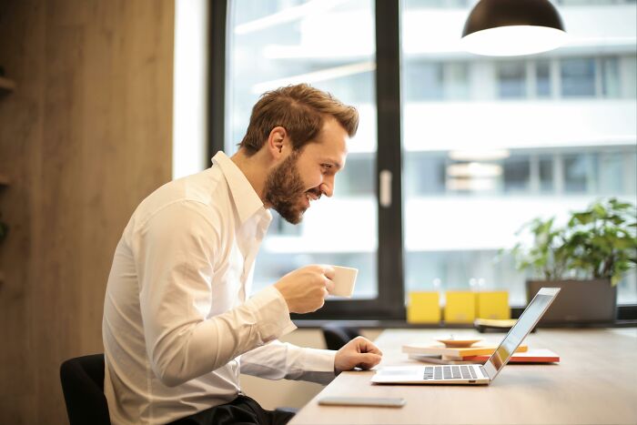 Man at desk looking at laptop screen, holding a coffee cup, with window in background, illustrating an unethical life hack.