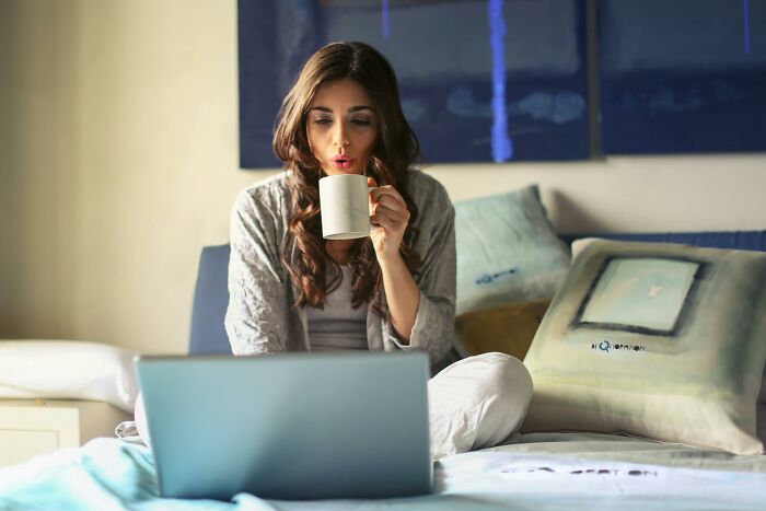 Woman sipping coffee while using a laptop on bed, reflecting on activities losing appeal with age.