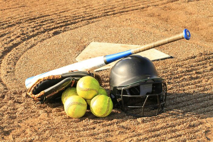 Softball gear on dirt field, featuring a bat, balls, glove, and helmet.