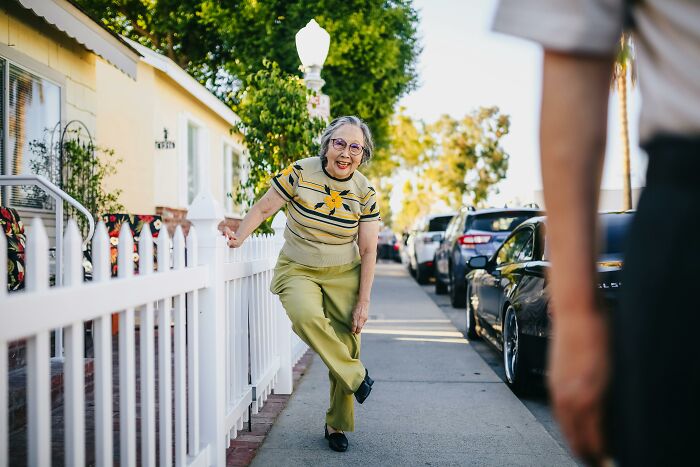 Older adult smiling while leaning on a fence in a colorful neighborhood street.