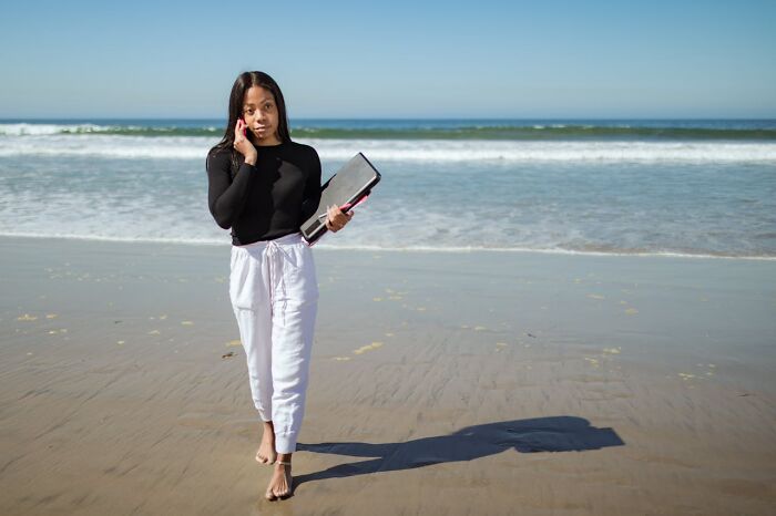 Woman walking on a beach, holding a folder and talking on a phone, reflecting on social norms.