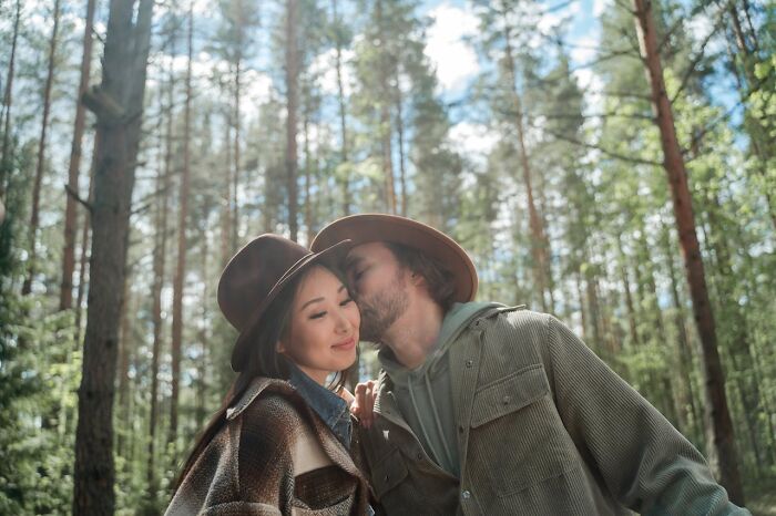 A man kisses a woman's cheek in a wooded area, both wearing hats, illustrating non-sexual acts that turn women on.