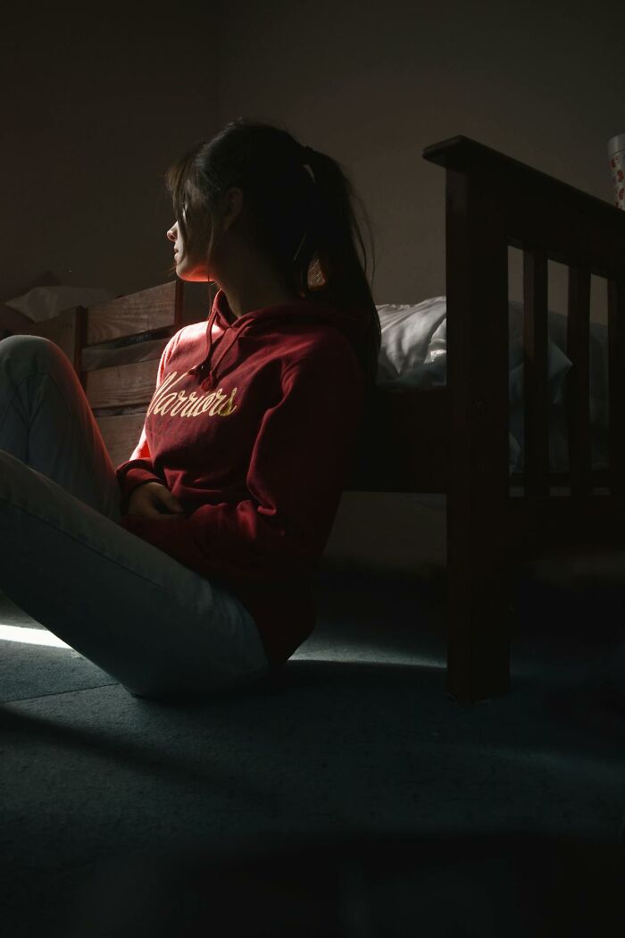 Young woman in a red hoodie sitting by a bed in a dark room, illustrating the impact of harmful social norms.