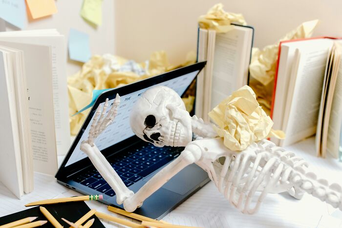 Skeleton slumped over a laptop surrounded by open books and crumpled papers, symbolizing millennial adulthood struggles.