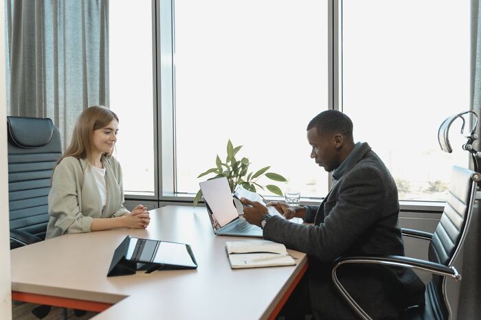 Two professionals at a desk with laptops, discussing changes to social norms in a modern office setting.