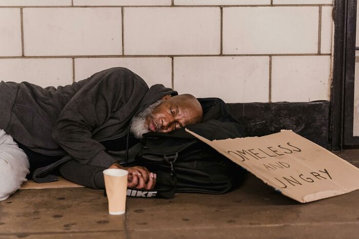 Homeless man sleeping on the ground with a sign and cup, illustrating unethical life hack concepts.