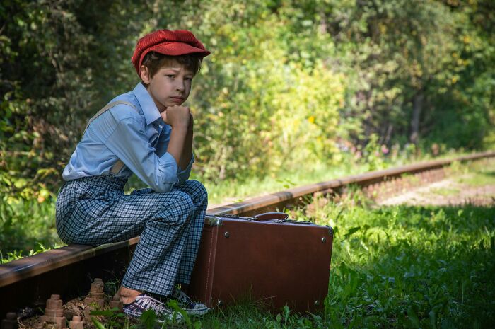 A boy in a red hat and plaid pants sits pensively by train tracks with a suitcase, evoking a mysterious atmosphere.