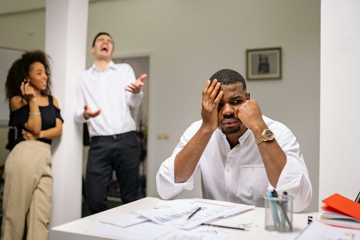 Man looks frustrated at desk while colleagues laugh, illustrating a savage job exit.