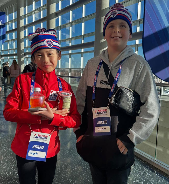 Adorable child ice skating duo wearing matching hats and badges, smiling indoors.