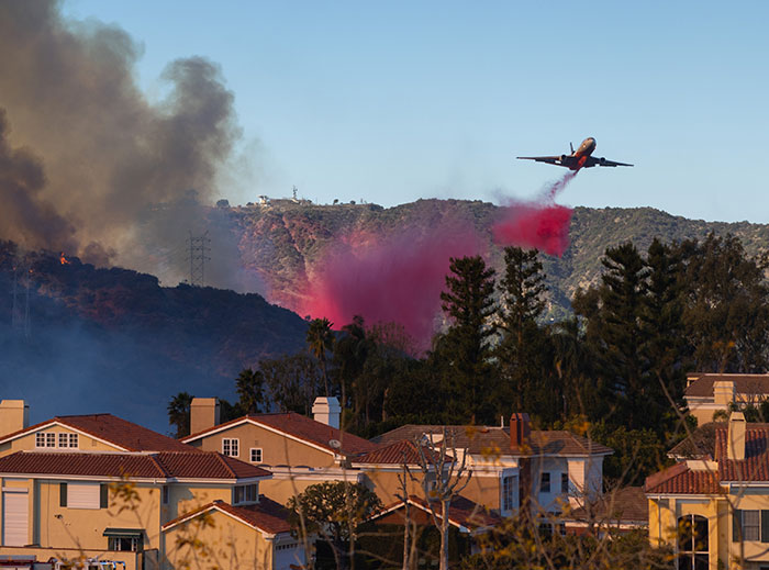 A firefighting plane drops fire retardant over hills near homes, combating wildfires and aiding private firefighters' efforts.