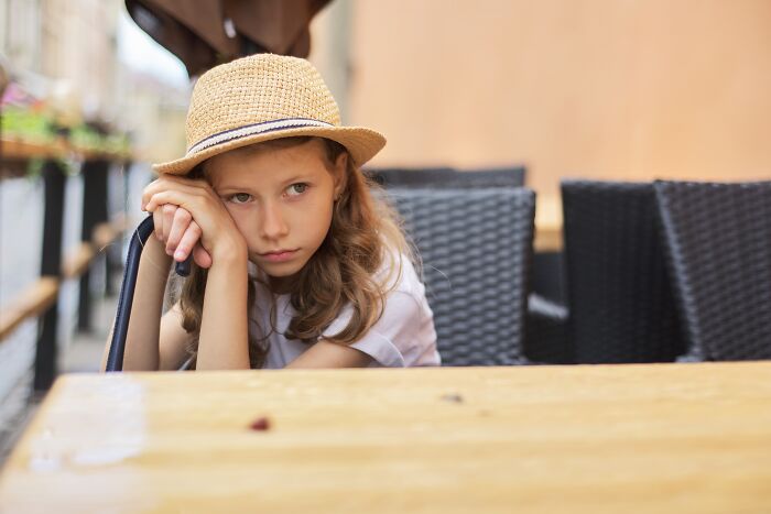 A sad child in a straw hat, seated at an outdoor café, symbolizing the impact of a horrible parent.