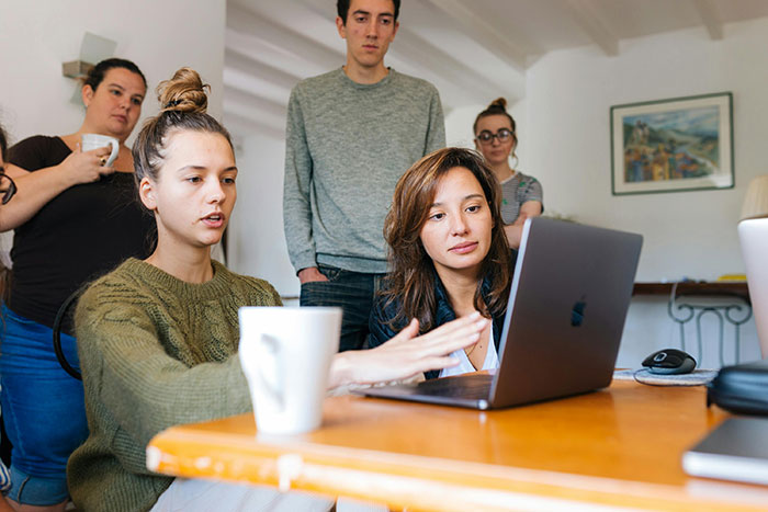 Group discussion around a laptop, two women focused, others watching, in a casual work setting.