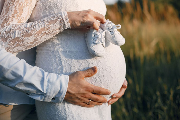 Pregnant woman in lace dress holding baby shoes, hands supporting her belly, outdoors.