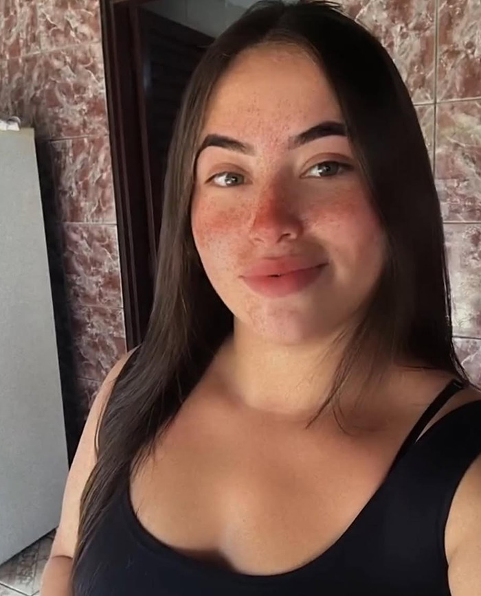 Young woman in a black top, smiling softly, with long brown hair and freckles, indoors.