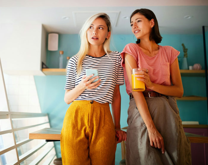 Two women casually chatting in a vibrant café, holding drinks and wearing stylish outfits.