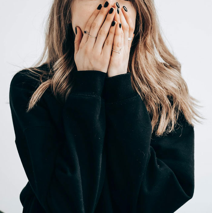 Woman covering her face with hands, wearing a black sweater, conveying a sense of embarrassment or shock.