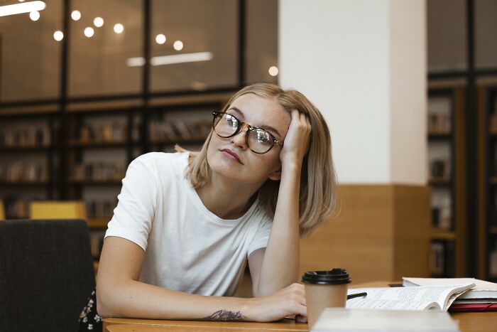 Millennial woman in a library, with glasses, looking thoughtful, sitting next to a coffee cup and open book.