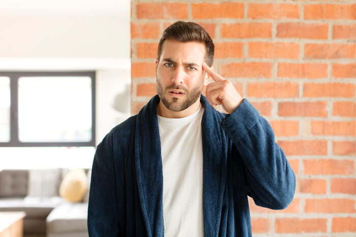 Man in a robe looking curious and pointing at his temple, demonstrating psychological tricks indoors.