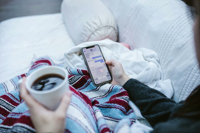 Person holding a phone and a cup of coffee, texting on a cozy couch, suggesting psychological tricks in communication.