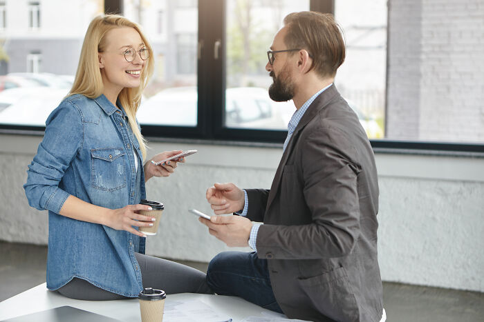 Two people chatting and smiling indoors, holding coffee cups and phones, illustrating psychological tricks in social interactions.