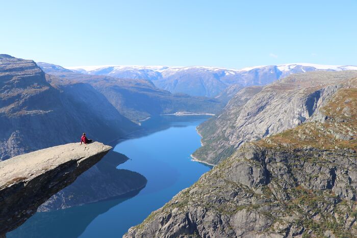 A person sits on a rock ledge overlooking a scenic fjord surrounded by mountains, evoking Tuscany Region in Italy's beauty.