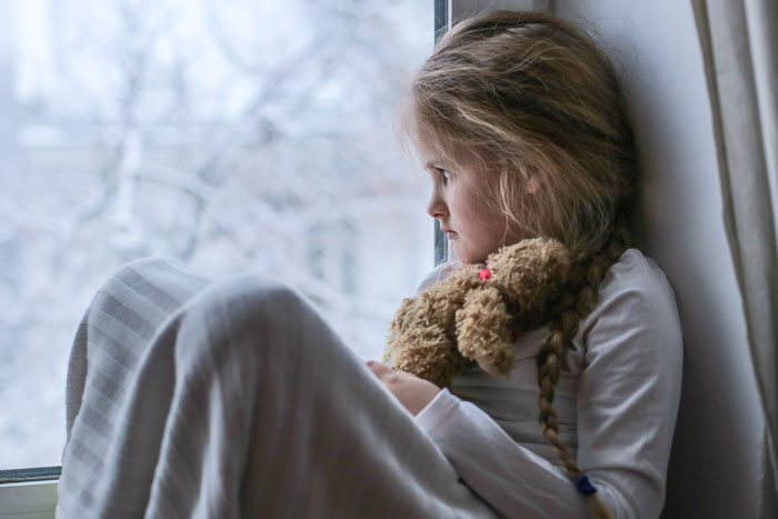 Young girl with a teddy bear gazing out a window, conveying emotions tied to family and hobbies balance.